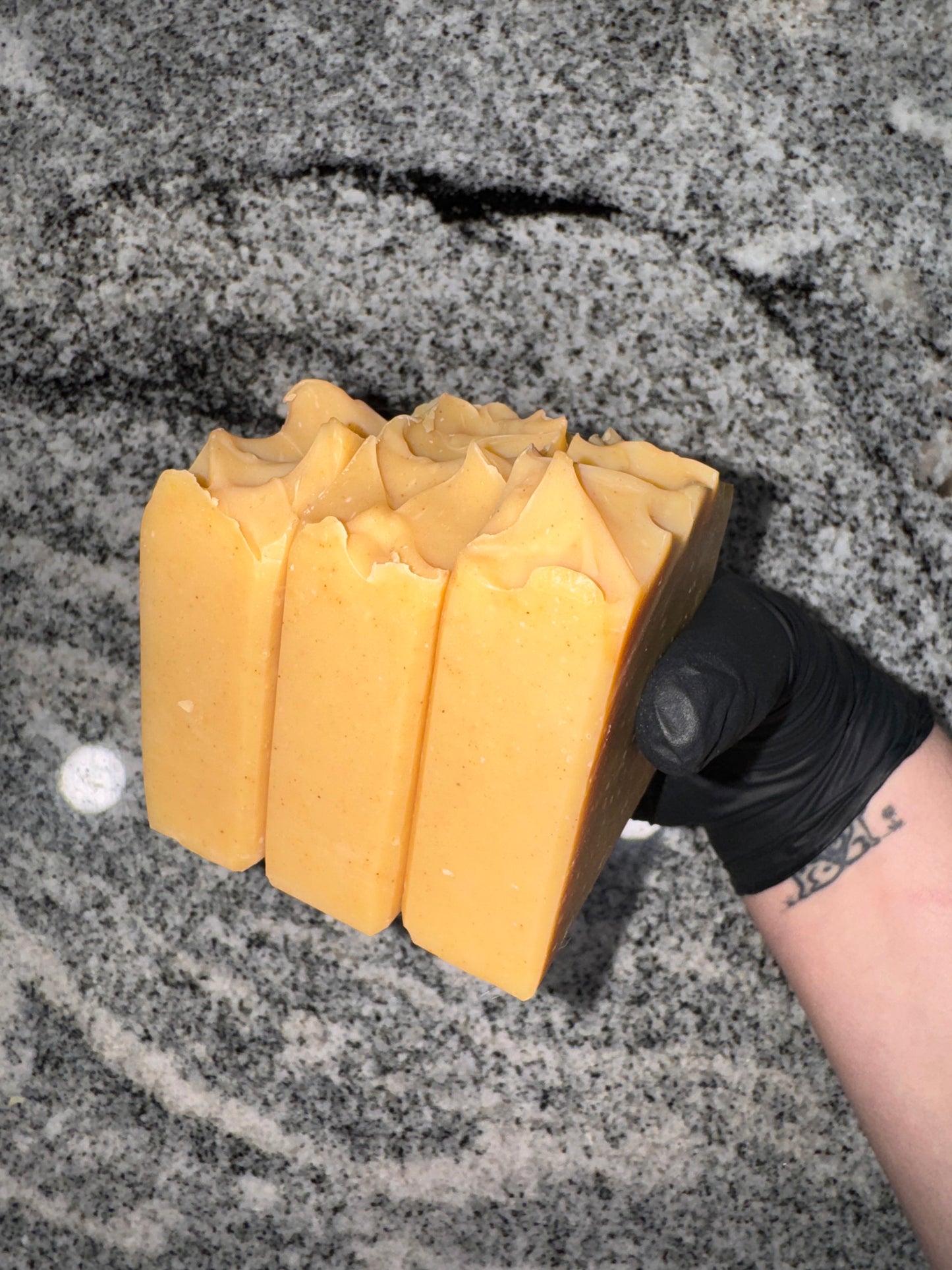 A gloved hand holds four orange soap bars with wavy tops against a speckled gray countertop. The mood is clean and artisanal.