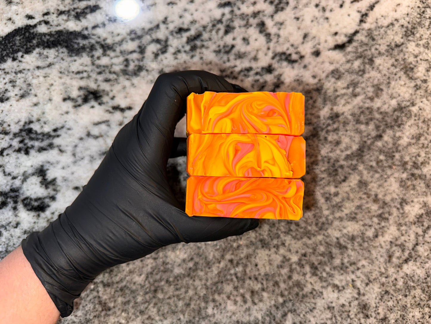 Three bright pink, orange, and yellow soap bars held by a gloved hand with a granite background.