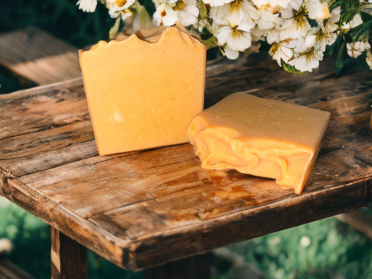 Two bars of textured orange soap on rustic wooden table with white flowers. The scene is natural and inviting, evoking a sense of freshness.