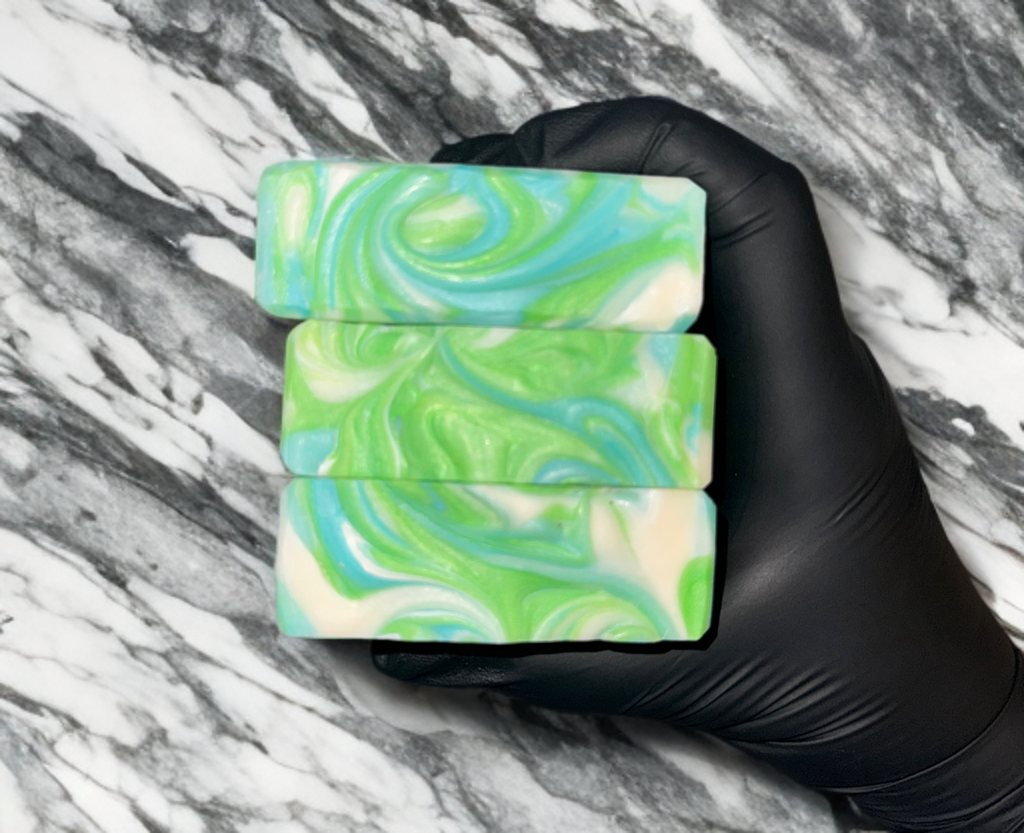 Three green, blue, and white swirled soap bars held by a hand wearing a black glove on a marble background