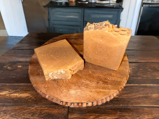 Two blocks of handmade soap sit on a wooden tray. One is upright with textured top, the other lies flat. The rustic setting adds a warm tone.