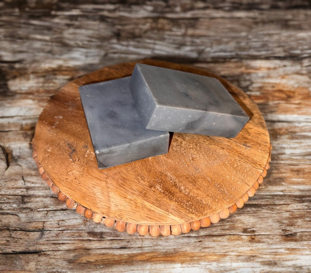 Two rectangular gray soap bars on a round wooden tray, set against a rustic wooden background, conveying a natural and earthy tone.