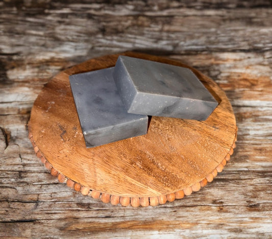Two rectangular gray soap bars on a round wooden tray, set against a rustic wooden background, conveying a natural and earthy tone.