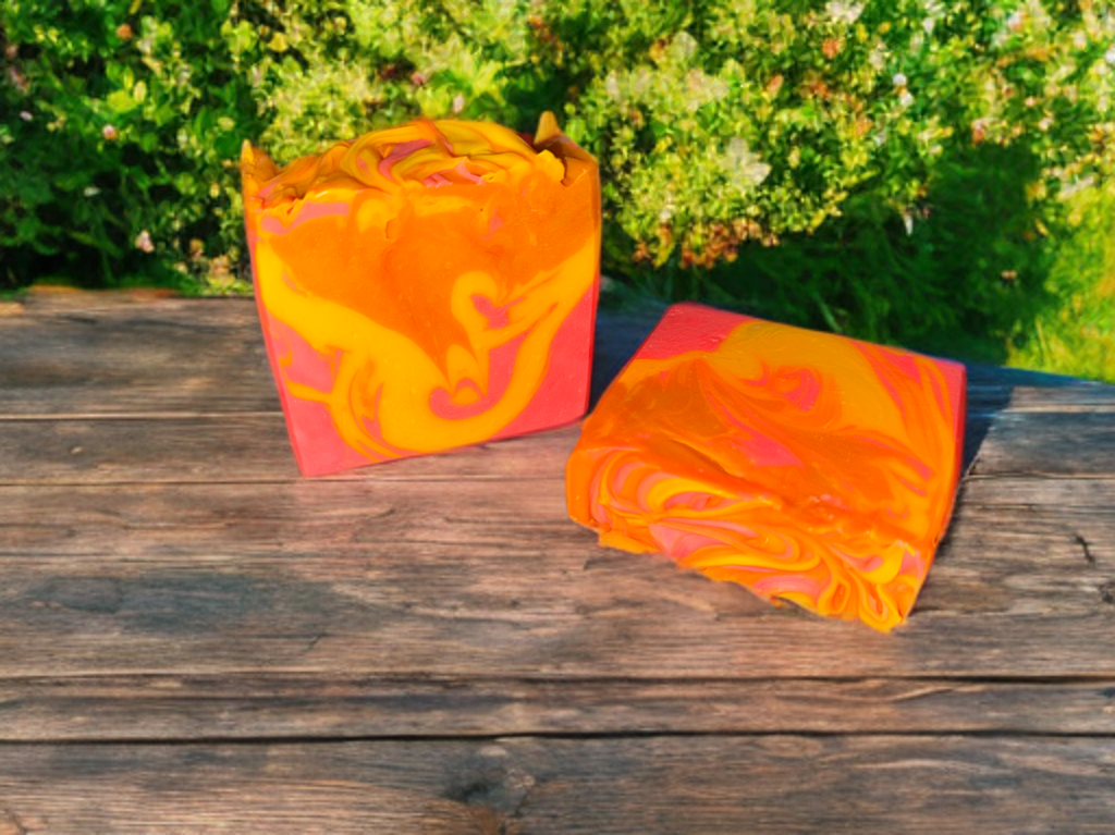 Two pieces of orange, pink, and yellow swirled soap bars on a wooden surface with greenery in the background.