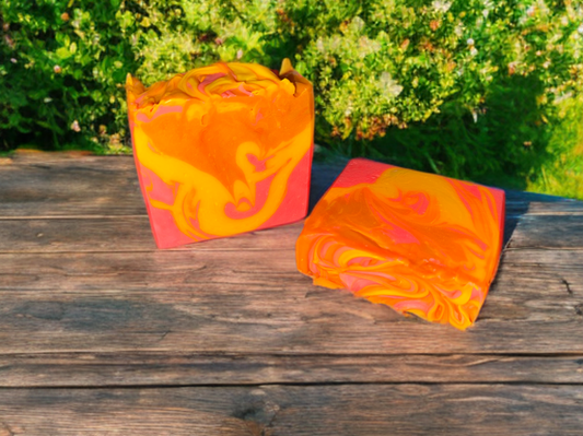 Two pieces of orange, pink, and yellow swirled soap bars on a wooden surface with greenery in the background.