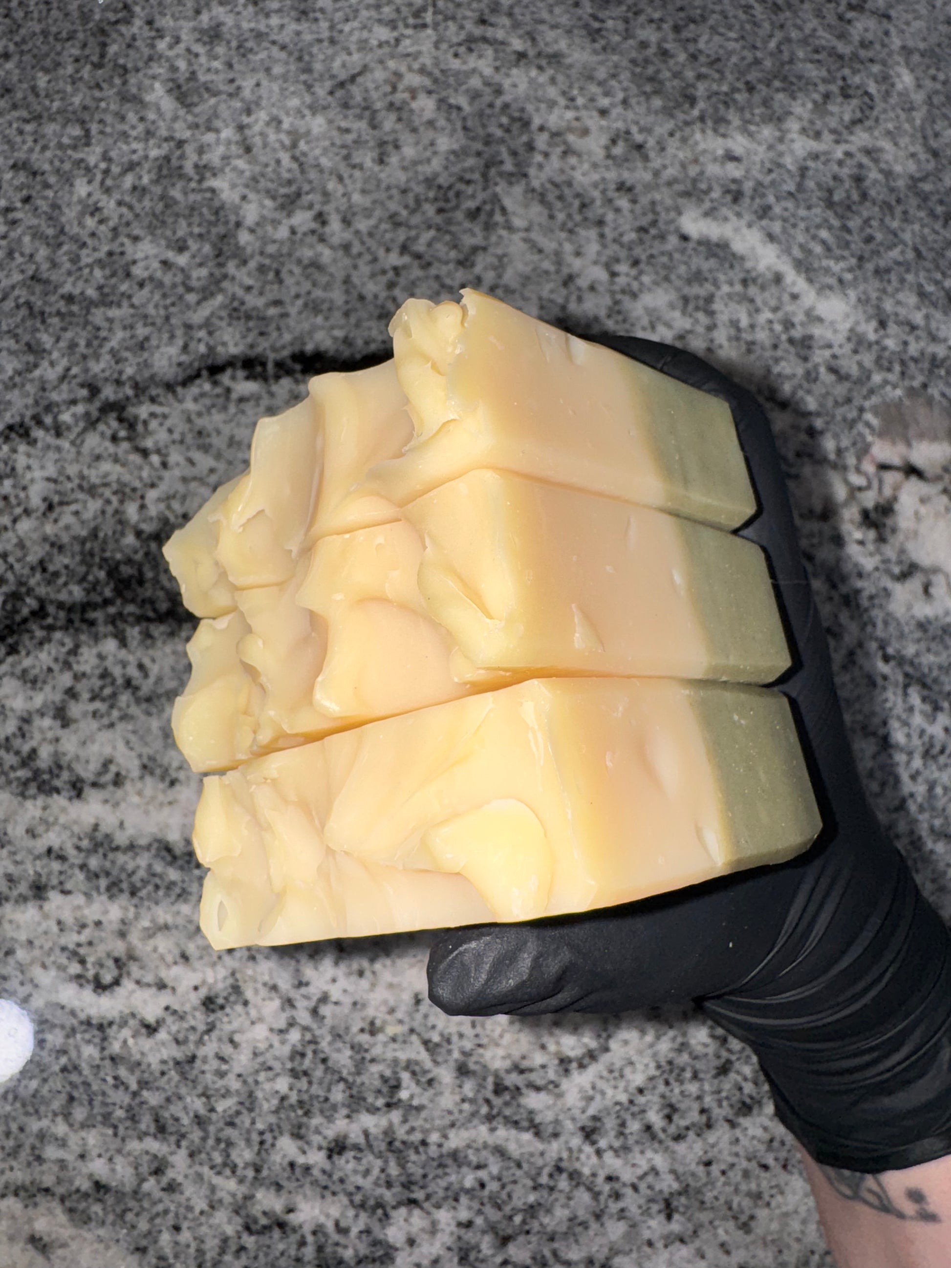 A gloved hand holds three handmade soap bars with swirled textures on a speckled gray countertop. The soap features soft cream and light green hues.