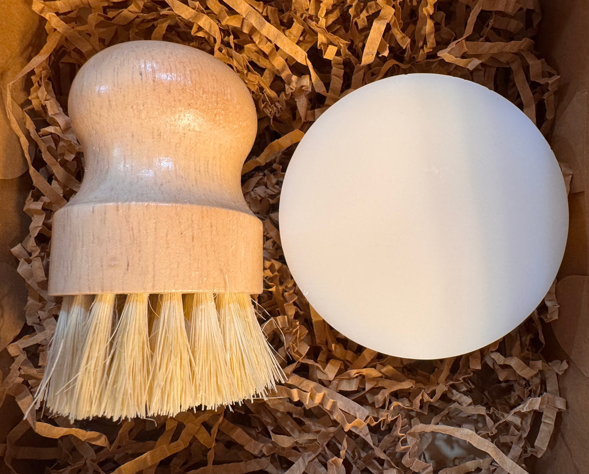 A natural wood-handled brush with pale bristles and a round white soap bar nestled in brown shredded-paper packaging, suggesting a simple, eco-friendly dish soap set.