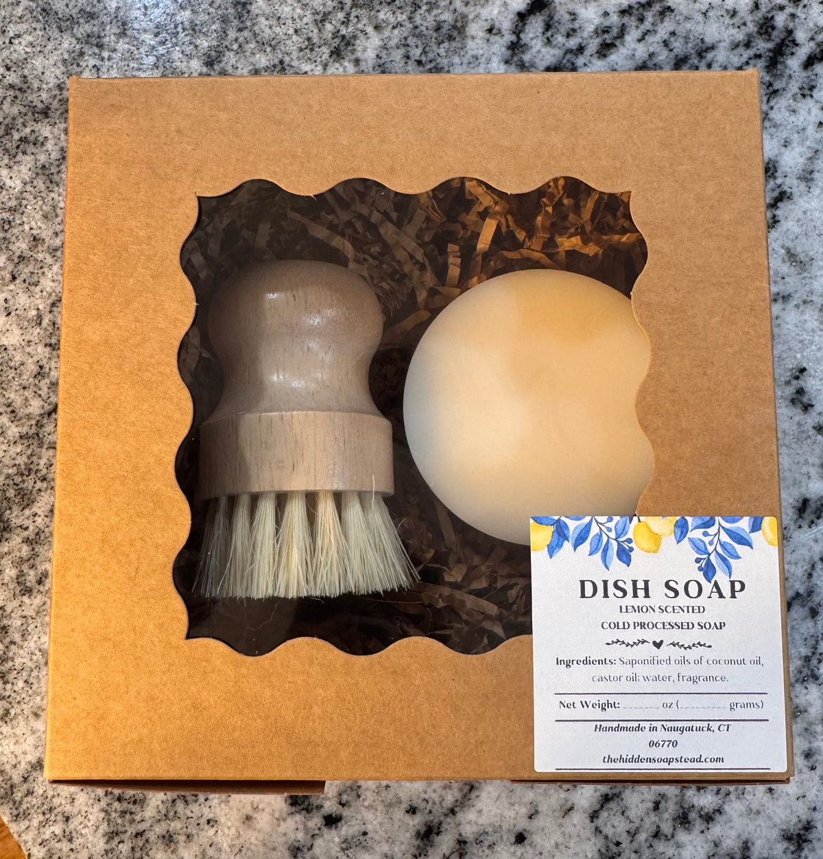 A kraft gift box with a scalloped window displays a wooden dish brush and round lemon-scented dish soap, with a blue-and-white label on a granite surface.